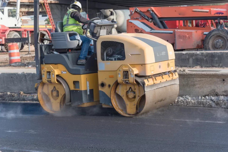A man operates a yellow roller to smooth out fresh pavement on a road.